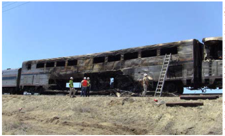 A burned out passenger train still on the track with investigators examining the wreckage.