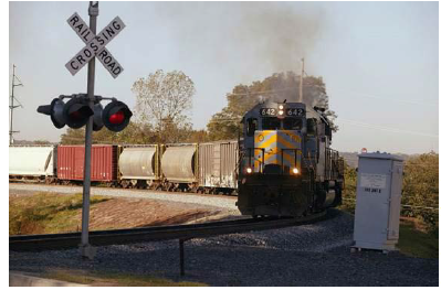 A train moving along a curve on the approach to a highway-railway grade crossing, where a pole-mounted crossbuck and red flashing lights warn oncoming traffic about the approaching train.