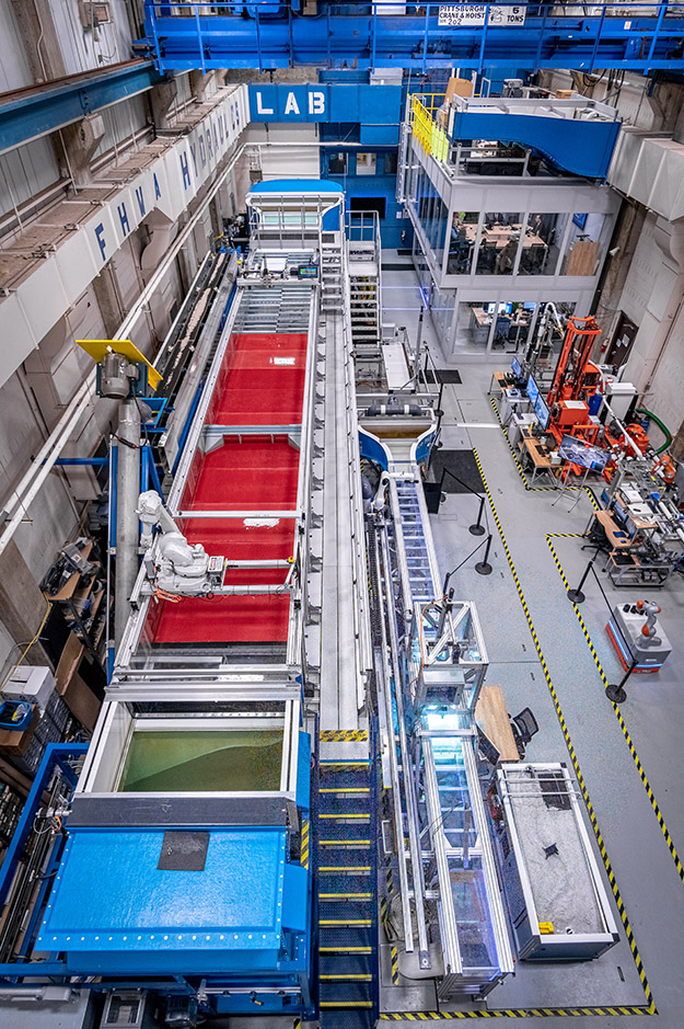 "The photo shows a birds-eye view of the FHWA Hydraulics Laboratory. The tilting flume is on the left. Next to the tilting flume is the computational fluid dynamics (CFD) station, ex situ scour testing device (ESTD), in situ scour testing lab sump device, and the force balance flume (from top to bottom). From top-to-bottom on the right: soil sample preparation station, presentation station, and the fish passage culvert flume."