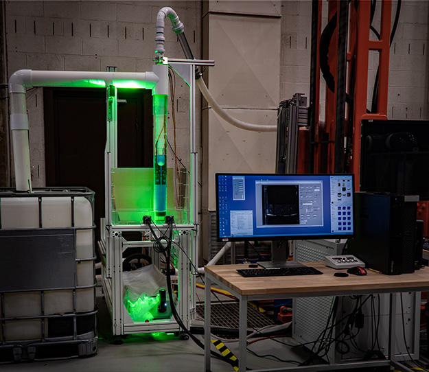 "the laboratory ISTD erosion head in clear piping illuminated by the laser, with two cameras mounted in front. In the foreground is the computer controlling the experiment."