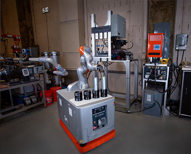 "the arm of an industrial robot reaching out towards a Shelby tube soil sample in preparation for loading it into the soil compaction station located behind the robot."