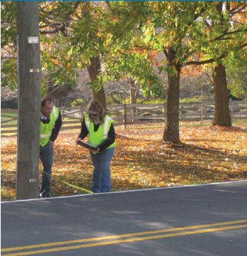 "A man and woman in retroreflective clothing measuring the distance from a utility pole to the edge of a raised roadway in a rural area and recording the data on a clipboard."