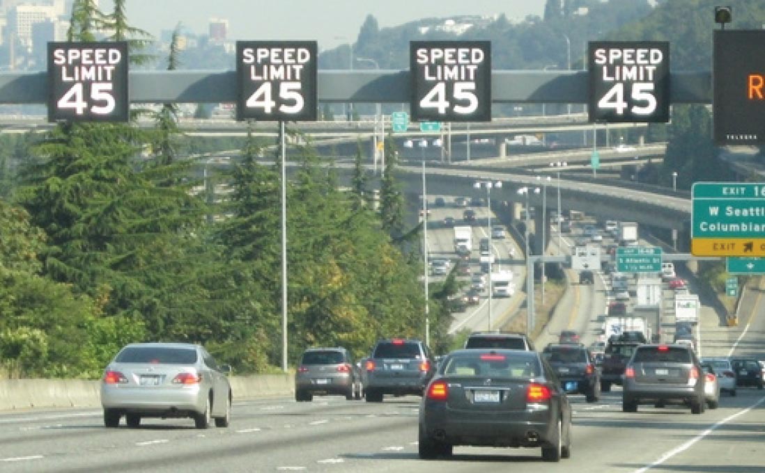 Four freeway lanes in the same direction. Over each travel lane is a variable speed limit sign reading SPEED LIMIT 45. To the right of the overhead variable speed limit signs is the left edge of an overhead variable message board sign. Three of the four travel lanes are congested, except for a restricted travel lane on the left.