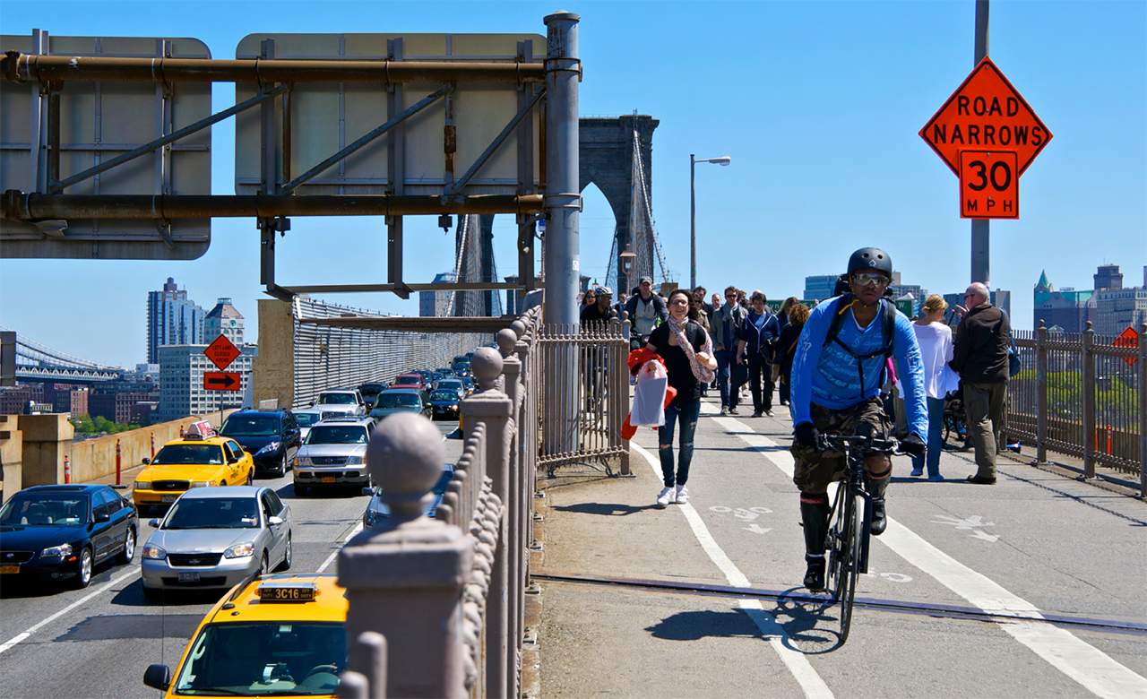 ""photo from Unit 2 of the Road Safety Fundamentals report showing cars, pedestrians, and a bicyclist crossing the Brookyn Bridge into Manhattan on a sunny day