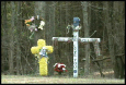 "This image shows a roadside memorial marking a fatal accident involving a teenage boy. The memorial includes two crosses, a wreath, flowers, and several personal mementos."