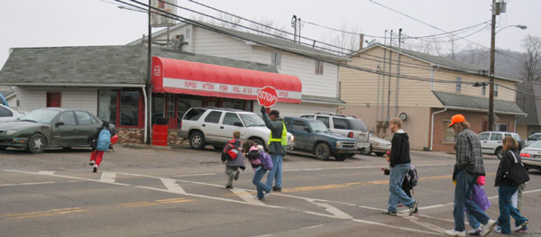 "Photo.  An adult school crossing guard stops traffic on a two-lane road though a rural community to allow children to safely cross the street."
