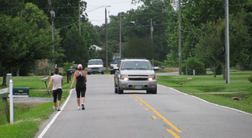 "Photo.  A rural two-lane  road with no shoulder .  Two pedestrians walk on the far left, facing traffic.  An on-coming vehicle has to encroach on the opposite travel lane to avoid the pedestrians."