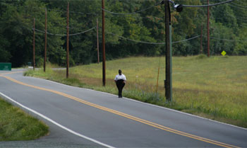 "Photo. A pedestrian walking on the right side of the road, the same direction as motorized traffic, on a road with no shoulder."
