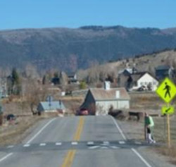 "Photo. A speed table, which is a raised of section of roadway – wider than a speed hump. It has a flat section on top that is wide enough to accommodate an entire car. The speed table in the photo is located at a crosswalk. In the photo, a pedestrian is begging to enter the crosswalk, which is signed and has pavement markings but is raised above the roadway."