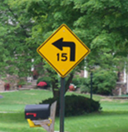 "alt="Photo. A yellow advisory sign with a black speed advisory of 15 MPH and curve warning. ""