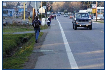 Photo of a staggered group of young people walking on the shoulder of a roadway in a rural town.