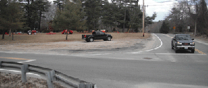 Photo at the intersection of Snake Hill Road at Chopmist Road (Route 102) showing a damaged guardrail in the bottom left corner. One truck is shown stopped at a stop line and another truck is shown parked along the side of the road.