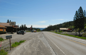 Photo of the eastbound view of intersection and parking/access locations just after the horizontal curve. A store is seen on the left hand side with some cars parked in a gravel covered parking lot. Another building across the street is on the right hand side in the distance with some trees and mountains in the distance.