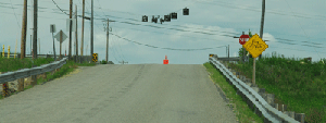 Photo of a northbound view on Dern road approaching the intersection with Highway 2. The intersection is on the crest of a steep vertical curve. On the hill approaching the intersection there are guardrails on each side. A bicycle warning sign is placed approaching the intersection and a stop sign with flashing beacons on a span wire are located at the intersection. Utility lines and poles are also seen crossing the intersection.