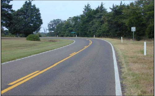 Photograph of a two-lane roadway with a curved section. The roadway has a white edge line on both side and double yellow centerlines. White delineators are mounted along the outside of the curve that match the color of the adjacent edge lines.