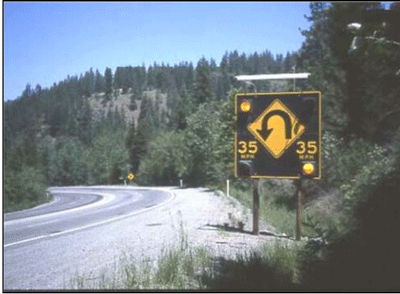 Photograph of a speed actuated sign installed within a two-lane curved section in Augusta, ME. The square sign has a black background. It has one flashing beacon on each corner and one hairpin curve sign in the middle. Above each of the flashing beacons on the bottom is the advisory speed sign of 35 mph.