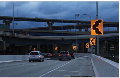 Photograph of a night view of sequential dynamic curve warning system, which are installed on the outside of a two-lane ramp. Solar powered flashing lights are embedded in the signs along the edges of the chevrons. The signs are larger than normal chevrons.