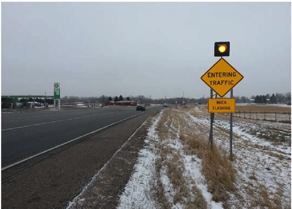 Photograph of a dynamic warning sign installed on the right side of a roadway. A flashing beacon is on top of the warning sign. The text on the warning sign reads Entering Traffic When Flashing.