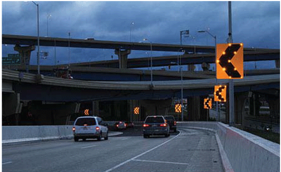 Photograph of a night view of sequential dynamic curve warning system, which is installed on the outside of a two-lane ramp. Solar powered flashing lights are embedded in the signs along the edges of the chevrons. The signs are larger than normal chevrons.