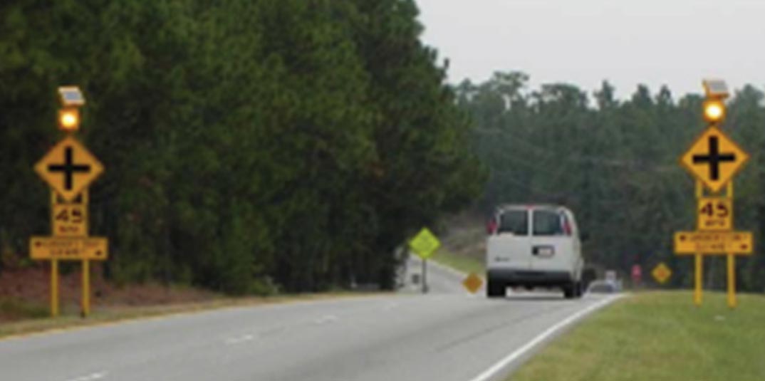 "Countermeasures present on the through approach of a two-way stop-controlled intersection. On both sides of the roadway, a sign assembly is present. From top to bottom, each sign assembly consists of an amber flashing beacon, a W2-1 intersection warning sign, a 45 MPH speed limit advisory plaque, and a yellow directional guidance sign, indicating the names of the cross street. Each sign assembly is supported by two metal posts covered in yellow reflective material. Additional intersection warning signs are visible in the background."