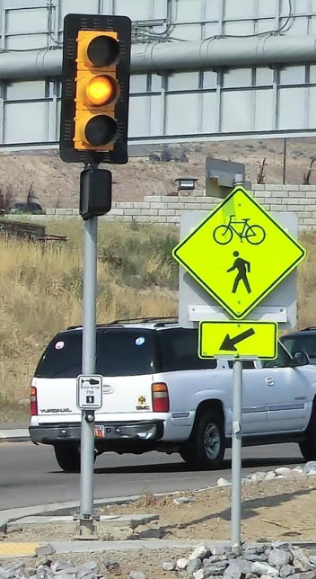 A traffic signal showing YELLOW at a trail crossing. The traffic signal is mounted with a black backplate onto a pole. Next to the pole supporting the traffic signal is a bicycle and pedestrian shared warning sign with supplemental directional arrow plaque. A white SUV is visible driving along the roadway in the background of the photograph.