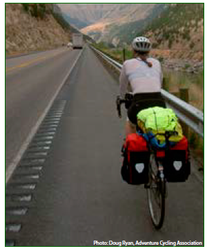 A bicyclist travelling on a wide shoulder that is edged on the left with rumble strips that contain gaps through which a cyclist may move without being forced to traverse the rumble strips themselves. Photo credit: Doug Ryan, Adventure Cycling Association.