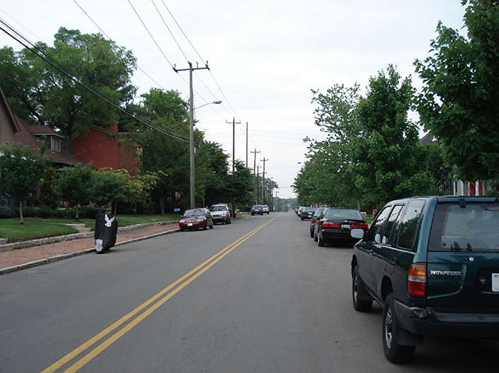 "Figure 3.19.3. On-Street Parking on a Collector Street. This figure contains a picture of a two lane street divided by a double yellow line. The picture is taken from the point of view of a passenger or driver in the right hand lane. Cars are parked parallel to the curbs in both lanes. Houses are visible on both sides and there are numerous trees."