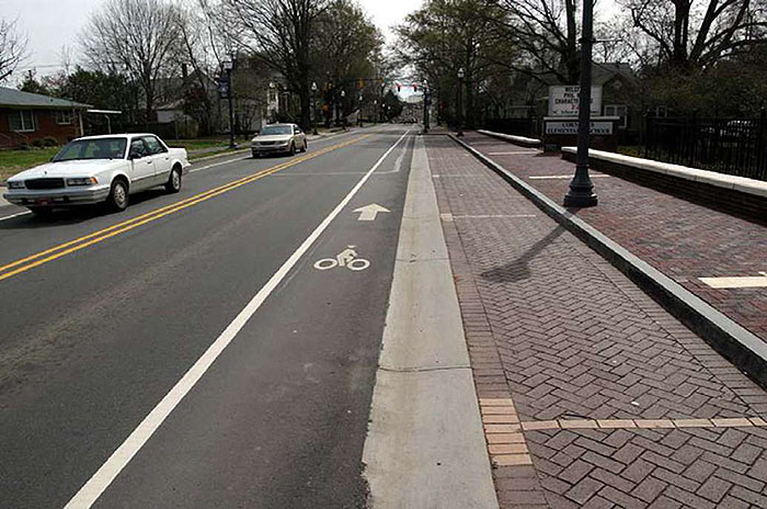 "Figure 3.19.4. On-Street Parking with Adjacent Bicycle Lane. This figure contains a photograph of a two lane street divided by a double yellow line. The picture is taken from the point of view of a car. A white car and a light colored car are driving in the left hand lane. The right lane is free of traffic. To the right of the right lane, there is a bicycle lane separated from the car lane by a solid white line. A bicycle icon and arrow indicate direction of travel and the purpose of the lane. To the right of that, there are parallel parking spaces with marked dividers. The spaces are done in brick. Further to the right is a sidewalk, also done in brick, and a lamp post. A low wall separates the sidewalk from a residential area. Trees are visible in the distance."