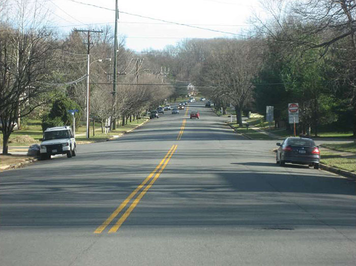 "Figure 3.20.2. Example Cross-Section Prior to Road Diet. This figure contains a photograph of a two lane road separated by a double yellow line. The lanes are wide and there are cars parked on each side. Trees and power poles line each side of the street."