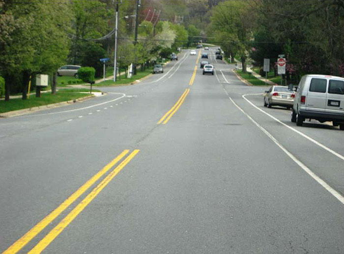 "Figure 3.20.3. Example Cross-Section with Road Diet. This figure contains a photograph of the same street in figure 3.20.2 after a Road Diet. The places where cars park are clearly delineated with solid white lines. There are also solid white lines just outside the parking areas to buffer the lanes. Cars are parked on both sides of the street."