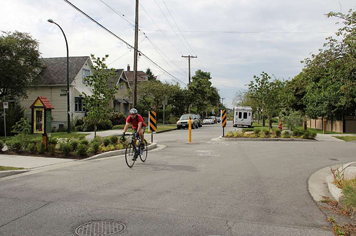 "Figure 3.21.3. Diagonal Diverter with Bicyclist. This figure contains a photograph taken from the leg of an intersection which curves to the right. Opposite this leg there is a section of road with parking on each side. A cyclist is riding through the channel, between the diverters. Yellow and black reflectors and a yellow post fill the channel to prevent motorized vehicle traffic. Houses line each side of the street. The diverters are lightly landscaped."