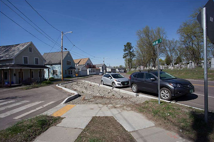 "Figure 3.22.1. Full Closure at Local Street Intersection with Collector. This figure contains a photograph of a former intersection which has been closed off by a set of concrete curbs. A channel has been left for drainage and the passage of bicycles. The leg which has been closed off, running top right to bottom left in the picture, is flanked by a row of houses. The leg which runs from mid-left to bottom right of the picture has parallel parking spots, filled by two compact cars. Sidewalks can be seen near the bottom of the picture and they both lead to a crosswalk which crosses the closed off street from right to left."