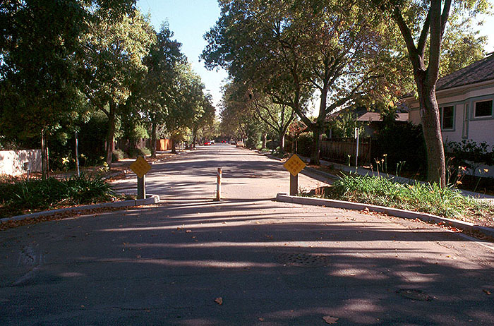 "Figure 3.22.2. Full Closure Located Midblock.This figure contains a photograph of a wide street in a residential area. The street has been narrowed by two curbs extensions landscaped with ornamental grasses. The closure consists of a diamond shaped yellow sign at the end of each curb extension and a post in the center of the channel. This allows for foot and bicycle traffic to pass through easily. There are a number of large trees and houses are visible along the right hand side of the street."