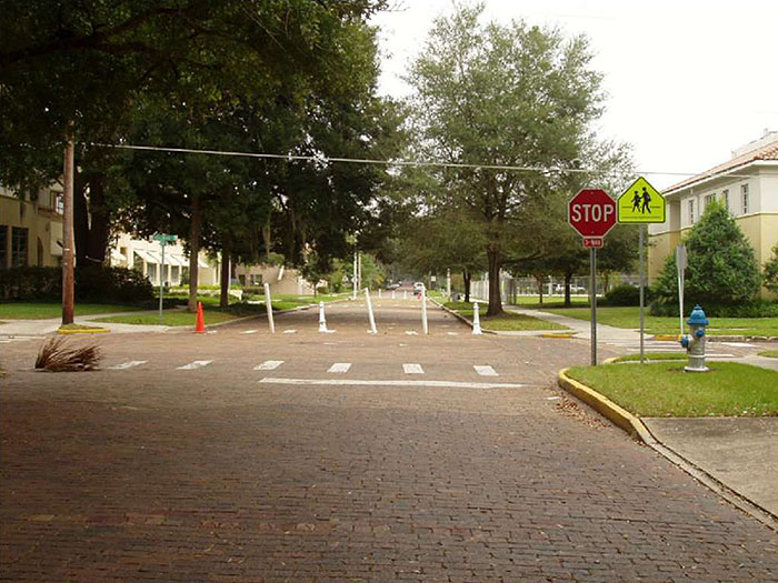 "Figure 3.22.3. Full Closure on One Leg of a Four-Leg Intersection. This figure contains a photograph of a four leg intersection taken from the point of view of a car in a leg at the bottom of the photo. A crosswalk crosses each leg of the intersection. The leg opposite the point of view camera is closed off by white posts. The right hand corner of the intersection has â€“ from right to left â€“ a blue fire hydrant, a school crossing sign, and a stop sign. Several trees are visible in the far leg and there is a house on the far right hand corner of the intersection, nearest the closure."