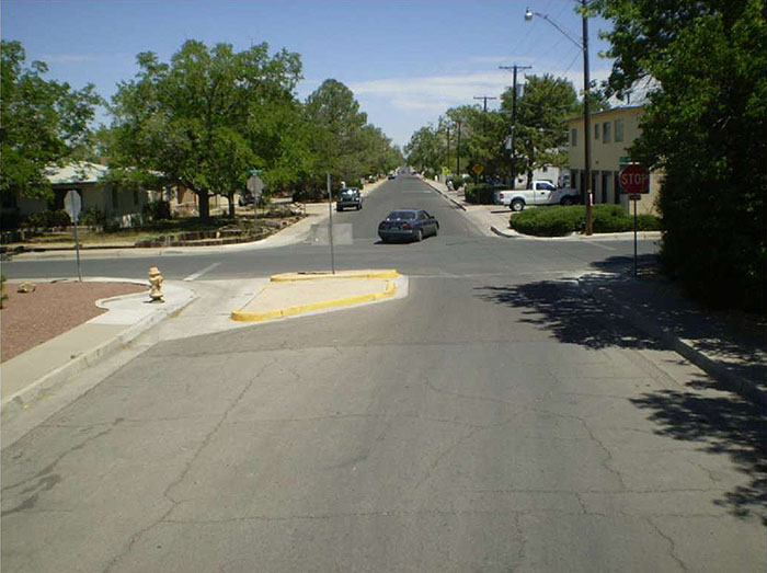 "Figure 3.23.2. Half Closure Blocking Entry to Side Street. This figure contains a photograph of an undivided two lane street running top to bottom of the photo with half closure blocking entry to a side street. A separated curb extension cuts the left lane off from any oncoming traffic. A gutter between the curb and extension allows for drainage and the passage of bicycle traffic. A yellow fire hydrant is visible on the curb near the extension. A car can be seen turning from the left leg of the intersection onto the top leg. Trees and houses line the far leg. There are stop signs at each corner."