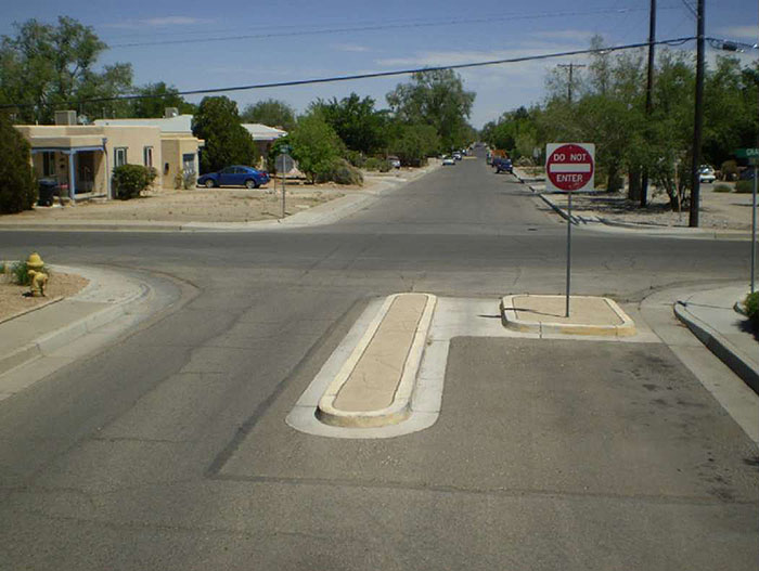 "Figure 3.23.3. Half Closure Blocking Exit from Side Street. This figure contains a photograph of an intersection with half closure blocking exit from side street. The lower leg of the intersection has a square curb extension in the right lane and a small median island. A Do Not Enter sign is mounted on a signpost on the curb extension. This configuration blocks any exit from this side street. There is no traffic visible. Some street parking can be seen in the distance on the top leg."