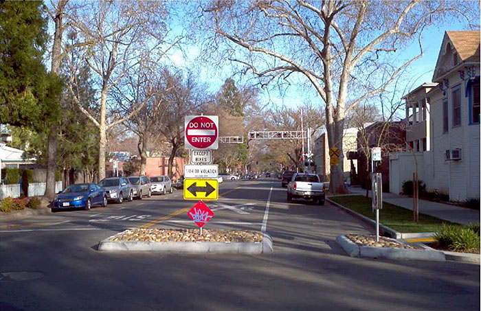 "Figure 3.23.4. Half Closure Blocking Entry to Side Street with Bicycle Cut-Through. This figure contains a photograph of one leg of an intersection. The left lane uses a curb extension and a median island to stop any traffic from turning onto it. The extension and island are filled with stone. Several signs, including "Do Not Enter", "Except Bikes", and a double headed arrow pointing left to right are mounted on a single signpost on the island. The separation between extension and median allows for bike traffic. A truck is parallel parked on the right hand side. A railroad crossing is visible in the distance. Cars are also parallel parked on the left lane. Houses and trees line both sides of the street."