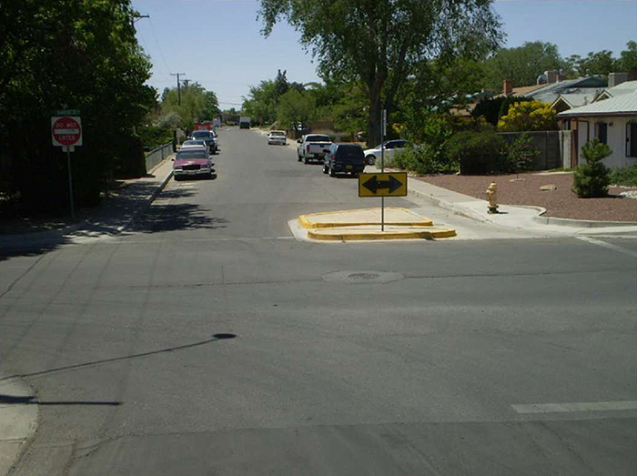 "Figure 3.23.5. Half Closure Allowing Exit from Side Street. This figure contains a photograph of the center of an intersection and the far leg. A curb extension with a cut through for pedestrian traffic obstructs any traffic from entering a side street, allowing for only exit. A signpost holds a double headed black arrow. A "Do Not Enter" sign is on the corner opposite the extension. Cars are parallel parked on either side of the street. A fire hydrant and a house can be seen on the same corner with the extension."