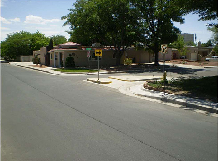 "Figure 3.23.6. Example Signage for a Half Closure Blocking Entry to a Side Street. This figure contains a photograph of a broad street with a side street intersecting it. A curb extension allows for foot and bicycle traffic but prevents any vehicle traffic from entering the side street. A "No Right Turn" sign can be seen on the right hand side of the street near the side street. A double headed arrow is attached to a signpost on the extension. Another "Do Not Enter" sign and a street sign can be seen on the corner opposite the extension. There is a house and trees beyond those signs."