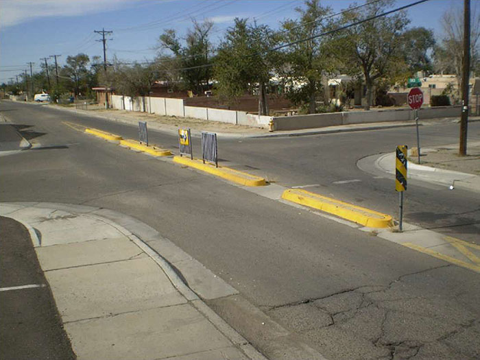 "Figure 3.24.4. Narrow Median Barrier with Cut-Throughs. This figure contains a photograph focusing on one street running from bottom right to top left of the picture. A narrow concrete median barrier blocks motor vehicle traffic from a cross street. Numerous cut-throughs allow for bicycles to pass. Yellow markers and turn only signs show motor vehicle traffic how to proceed. There are trees in a lot opposite the corner where the picture was taken from."