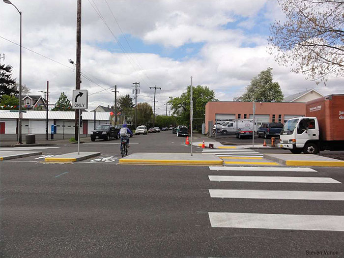 "Figure 3.24.5. Median Barrier with Pedestrian Refuge and Bicycle Cut-Through. This figure contains a photograph taken from a cross street. A wide median barrier runs from left to right. On the left hand side there are two cut throughs marked for bicycle traffic with white bicycle icons painted on the pavement. A left turn only sign is mounted on the small island formed by these cuts. A pedestrian refuge on the right hand side of the picture is formed where the crosswalk crosses over to the far side of the street, providing a place for foot traffic to wait. A truck can be seen in the lane opposite the median and industrial buildings can be seen in the lots opposite the street."