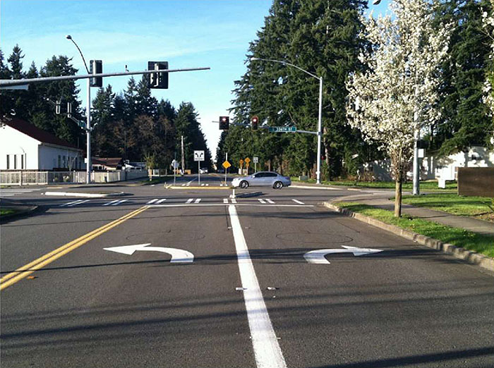 "Figure 3.24.6. Forced Turn Island Blocking Side Street Through Movements and Allowing All Turns. This figure contains a photograph which focuses on a left turn only lane and a right turn only lane on the near side of a cross street. The far side is blocked by a median barrier with a sign indicating that both left and right turns can be made here. A car is moving through the intersection from right to left. There are stop lights on arms visible on each leg of the intersection."