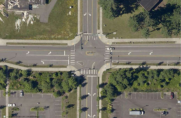 "Figure 3.24.7. Forced Turn Island Blocking Side Street Through Movements and Allowing All Turns â€“ Aerial. This figure contains an aerial photograph showing a complete intersection. The left, top, and bottom legs allow left and right turns. The top and bottom legs can't be entered from opposite lanes. Cross traffic is only allowed on the street running left to right. A collection of islands are used to manage the turns."