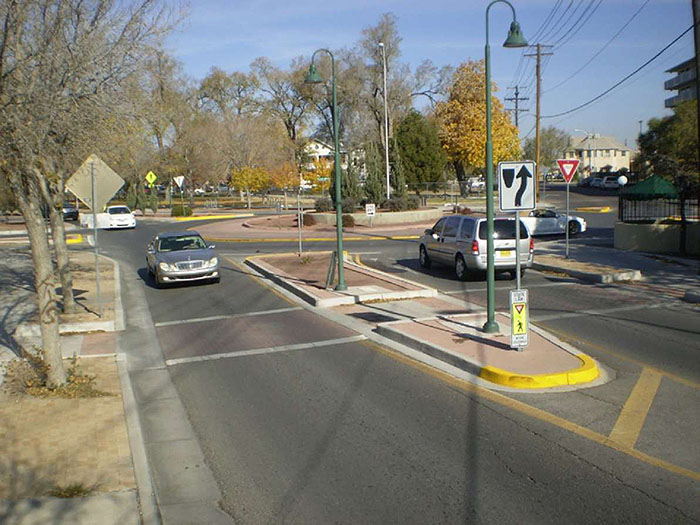 Figure 3.9.2. Roundabout with Urban Cross-Section (i.e., Curb and Gutter). This figure contains a photograph of an entrance/exit for a roundabout. A car enters it on the right hand side. It is separated from a car exiting the roundabout by a splitter island. Trees and other landscaping can be seen in the center of the circle.