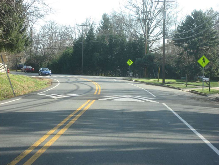 "Figure 3.10.3. Speed Hump Along Horizontal Curve. This figure contains a photograph of a curving two lane road divided by a double yellow line in a wooded area. Just before the curve a speed hump is in place a short distance from a cross walk. The crosswalk is indicated by green diamond signs on the right hand side of the road."