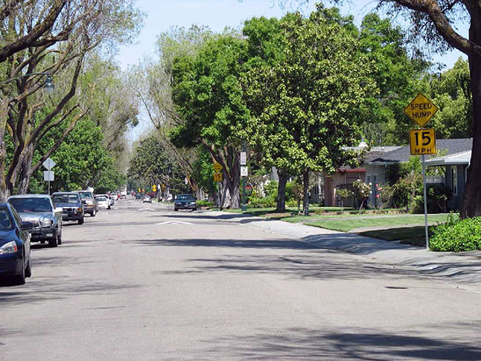 "Figure 3.10.5. Series of Speed Humps. This figure contains a photograph of an undivided street in a tree-lined residential area. A sign post with a yellow diamond speed hump sign and a rectangular 15 MPH speed limit sign is on the right hand side of the street. A series of speed humps is visible as the street vanishes into the distance."