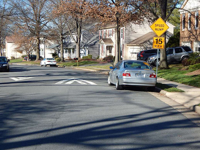 "Figure 3.10.6. On-Street Parking Adjacent to Speed Hump. This figure contains a photograph of a curved street in a residential area. A car is parked near the curb beside a signpost with a yellow diamond speed hump sign and a 15 MPH speed limit sign. Other cars can be seen parked on either side of the street."