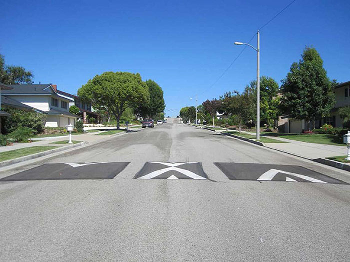 "Figure 3.11.1. Speed Cushion with Passage that Straddles Centerline. This figure contains a photograph of a tree lined undivided street in a residential area. Three speed cushions are visible, crossing the road from right to left. The right and left cushion are painted with wide white angular stripes indicating a speed cushion. The cushion which straddles the centerline has angular stripes on each side and is narrower than the other two."