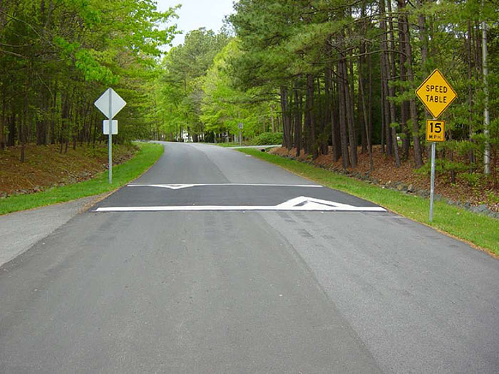 ""Figure 3.12.1. Typical Speed Table Application. This figure contains an unlined two lane road running through a wooded area. Running across the road is a speed table, a long raised area with a flat top. White angular striping marks the right of way on the speed table. Signposts on both approaches hold a diamond shaped yellow sign that says Speed Table over a rectangular yellow sign that says 15 MPH.