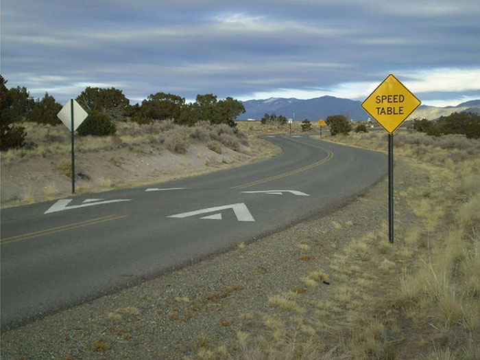 "Figure 3.12.3. Speed Table in a Series. This figure contains a photograph of a two lane road divided by double yellow lines curving from the bottom left of the picture to near its right edge and back to the center. A speed table is visible before the curve begins, striping painted on it to indicate right of way. On either side of the road, there is a signpost holding a diamond shaped yellow sign which says Speed Table. To the left of the road there is a hill with scrub brush. An additional speed table can be seen after the curve. The shoulder off of the right side is dirt and gravel"